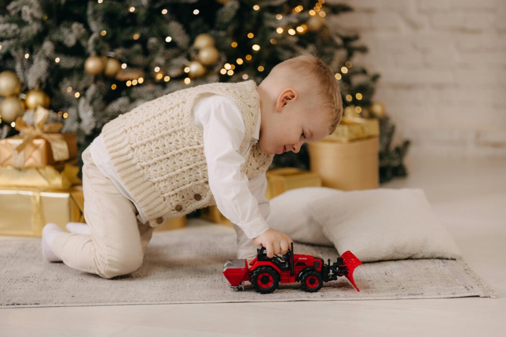 A young child plays with a toy tractor near a Christmas tree with gold decor.