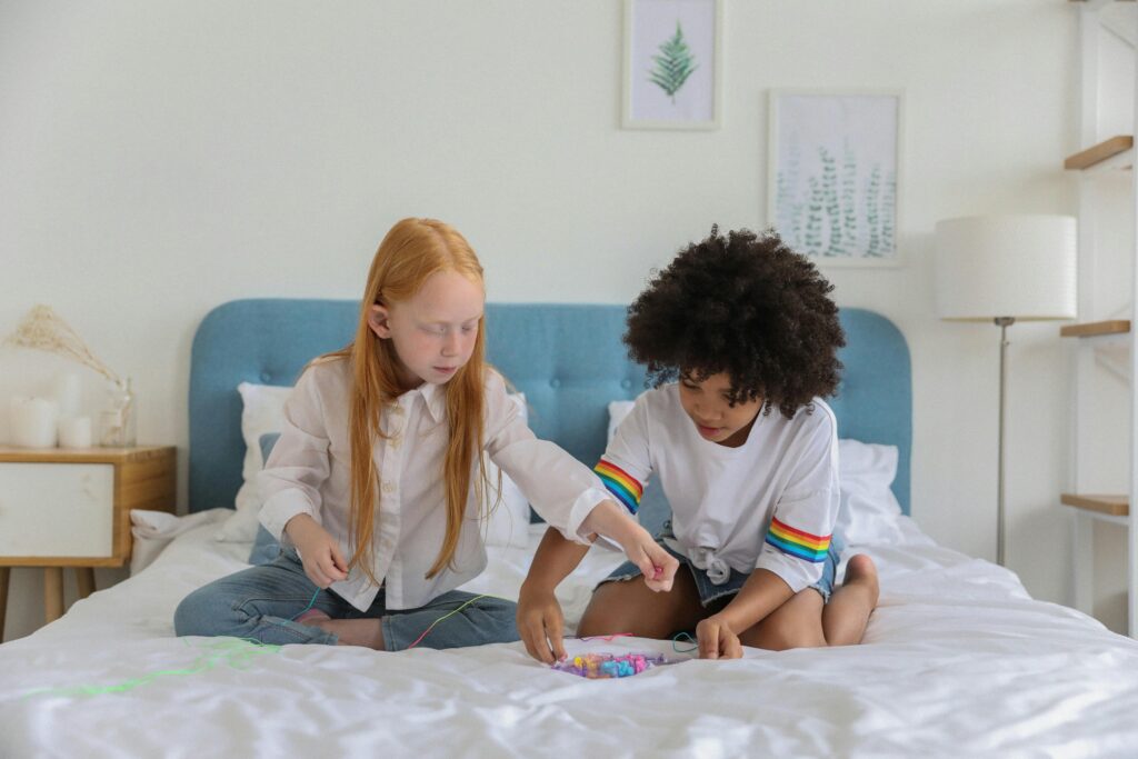 Two girls sitting on bed, interacting and creating friendship bracelet in a cozy bedroom setting.